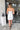 Full body back view of model wearing the Ice Queen Dress that has white fabric with a metallic silver floral pattern, a strapless neckline with pleated ruffle detailing, a lining, and a back zipper closure