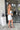 Full body side view of model wearing the Ice Queen Dress that has white fabric with a metallic silver floral pattern, a strapless neckline with pleated ruffle detailing, a lining, and a back zipper closure