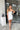 Full body front view of model wearing the Ice Queen Dress that has white fabric with a metallic silver floral pattern, a strapless neckline with pleated ruffle detailing, a lining, and a back zipper closure
