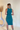 Back view of model wearing the Over The Pines Dress which features dark green ribbed fabric, slit on the left side hem, round neckline and sleeveless.