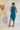 Back view of model wearing the Over The Pines Dress which features dark green ribbed fabric, slit on the left side hem, round neckline and sleeveless.