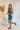 Side view of model wearing the Over The Pines Dress which features dark green ribbed fabric, slit on the left side hem, round neckline and sleeveless.