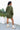 Full body back view of model wearing the One Way Ticket Romper, that features olive fabric, wood button closures, v neckline, two front pockets with tie details and balloon long sleeves with elastic wrists