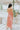Back view of the Coastal Crush Dress that features a coral colored material, a cowl neckline, a sleeveless design with thin adjustable straps, a side slit, and a midi length.