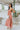 Side view of the Coastal Crush Dress that features a coral colored material, a cowl neckline, a sleeveless design with thin adjustable straps, a side slit, and a midi length.