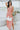 Frontal view of the Coastal Crush Dress that features a coral colored material, a cowl neckline, a sleeveless design with thin adjustable straps, a side slit, and a midi length.
