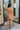Back view of the Something Like That Dress that features a salmon satin material, a cowl neckline with cups at the bust, a sleeveless design with thin adjustable straps, a waist cut-out, an elastic waistline, and a criss-cross strap back.
