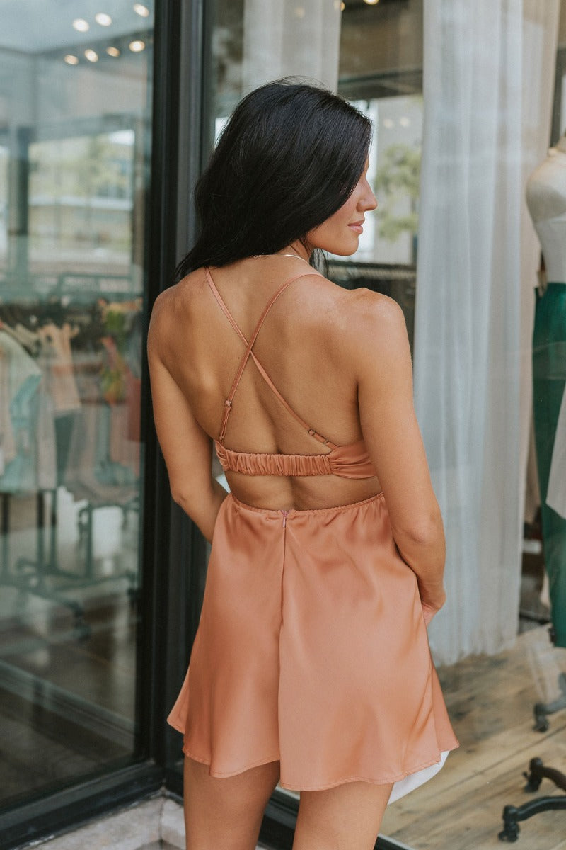 Back view of the Something Like That Dress that features a salmon satin material, a cowl neckline with cups at the bust, a sleeveless design with thin adjustable straps, a waist cut-out, an elastic waistline, and a criss-cross strap back.