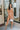 Frontal view of the Something Like That Dress that features a salmon satin material, a cowl neckline with cups at the bust, a sleeveless design with thin adjustable straps, a waist cut-out, an elastic waistline, and a criss-cross strap back.