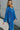 Side view of model wearing the Be You Pants which features royal blue pleated fabric, elastic waistband and flare pant with lettuce detail on the hem.