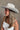 Side view of model wearing the Westside Ivory Cowgirl Hat which features cowboy shape, ivory suede fabric and rhinestone design on bottom of hat.