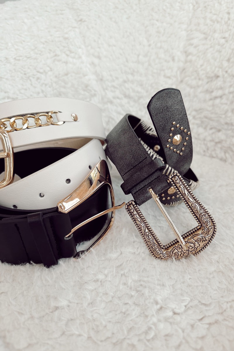 Front lay view of the Indy Black Western Adjustable Belt which features washed black fabric, ivory stitch details, gold medallion details and gold, western inspired, adjustable buckle. Belt pictured with other belts on display.