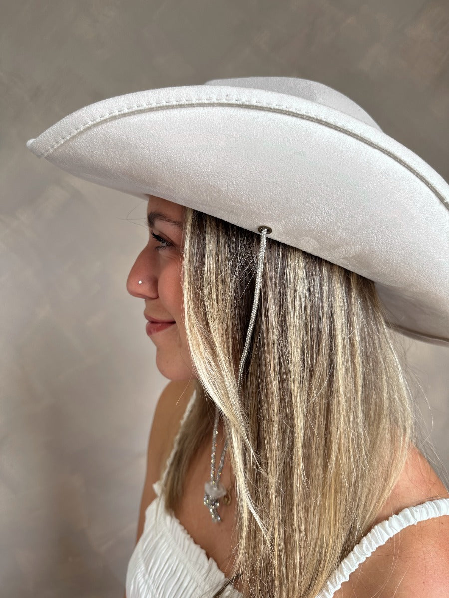 Side view of model wearing the Lainey Ivory Rhinestone String Cowgirl Hat which features cowboy shape, ivory suede fabric and rhinestone covered adjustable string.