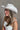 Side view of model wearing the Lainey Ivory Rhinestone String Cowgirl Hat which features cowboy shape, ivory suede fabric and rhinestone covered adjustable string.