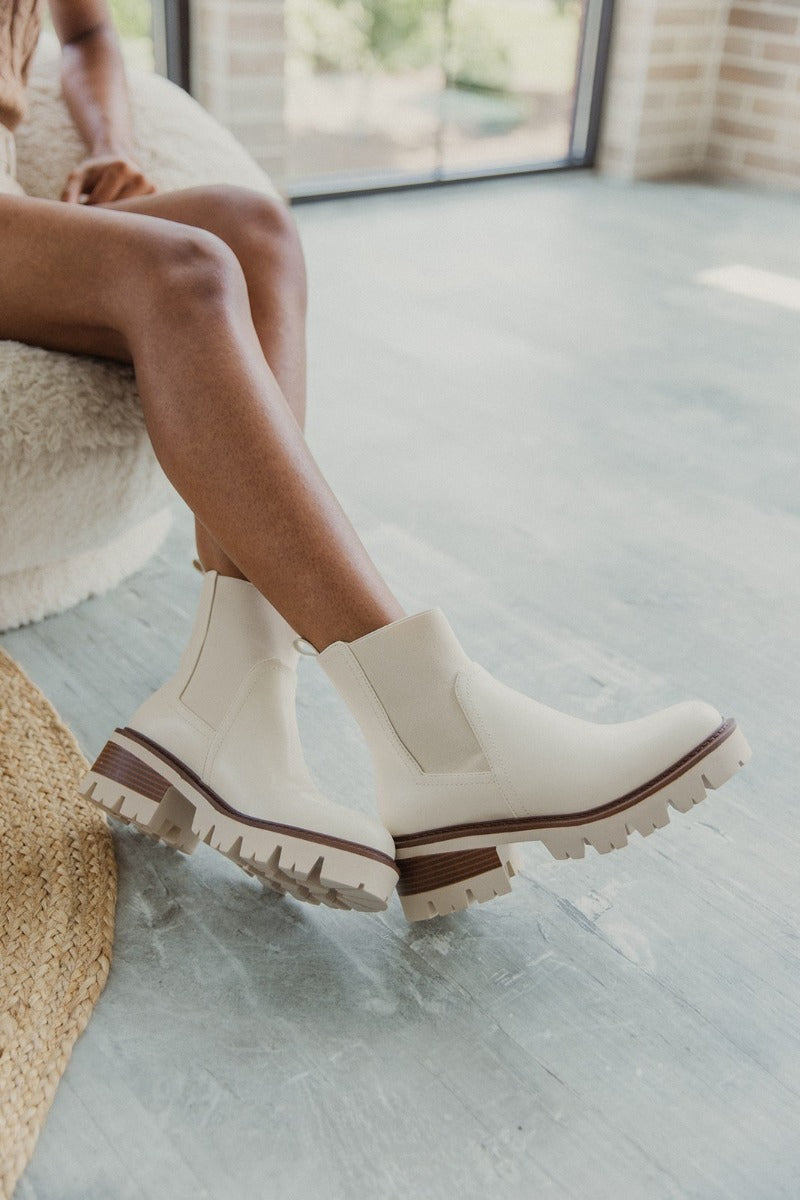 Front side view of model wearing the Ives Lug Chelsea Boot in Bone which features off white faux leather fabric, round toe, lug sole with brown color-tone details, contoured footbed and pull tab for easy slide-on.
