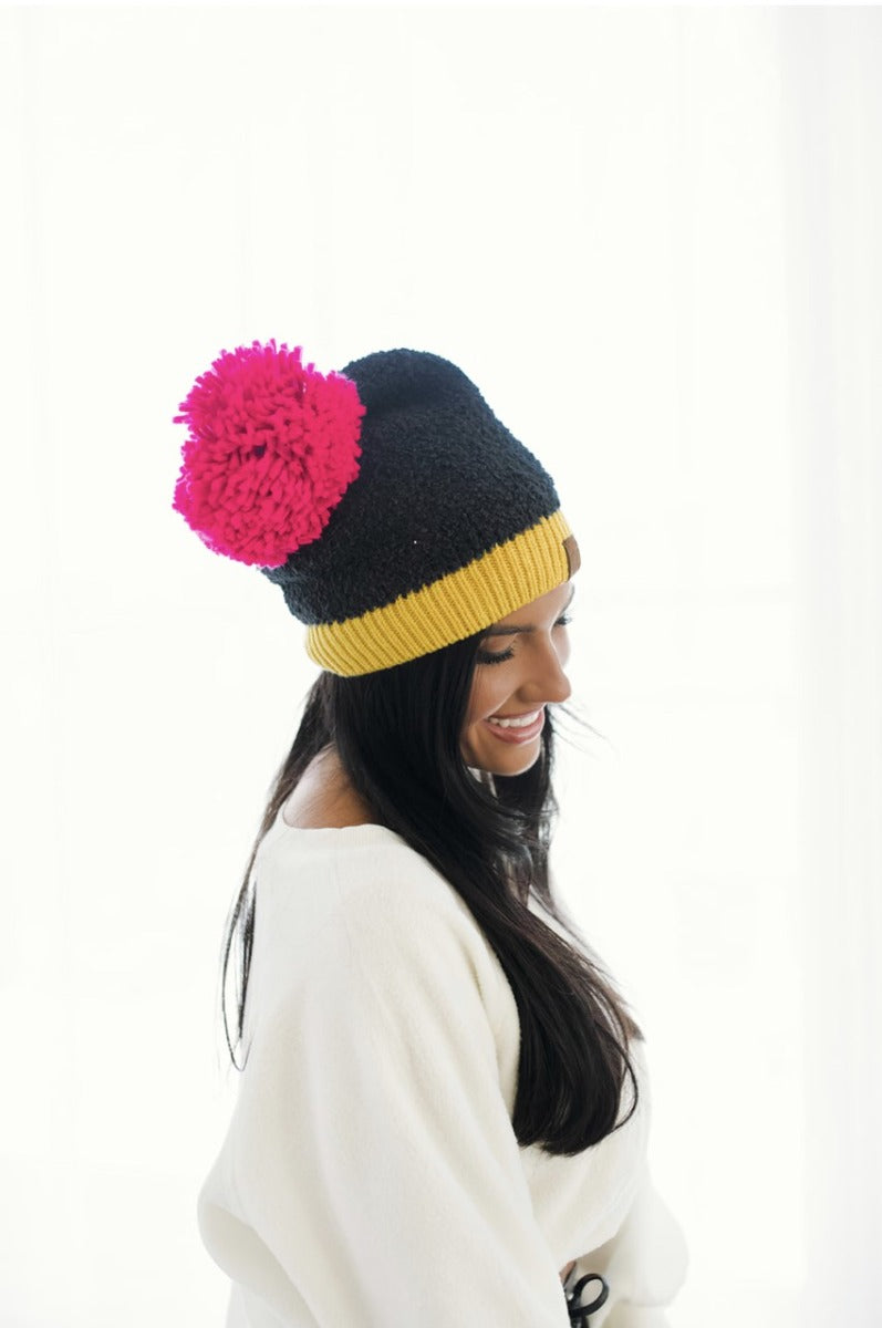 Side view of model wearing the Stay Cozy Beanie in Black, that features black sherpa fabric, yellow trim, and a pink pom pom.