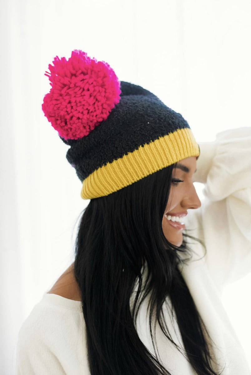 Side view of model wearing the Stay Cozy Beanie in Black, that features black sherpa fabric, yellow trim, and a pink pom pom.