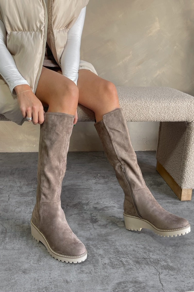 Front view of model wearing the Risky Boot in Dark Taupe which features dark taupe suede fabric, chunky lug platform sole, back pull tab, round toe and inside side monochrome zipper.